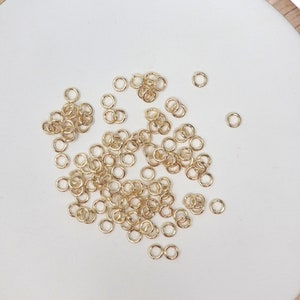 May include: A close-up of a white surface with a scattering of small, gold jump rings. The rings are arranged in a random pattern, with some overlapping.
