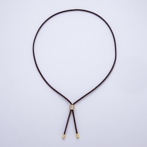 May include: A dark brown cord necklace with a gold-colored accent piece and gold-colored tips. The necklace is displayed against a white background. The cord forms a loop and then splits into two strands.