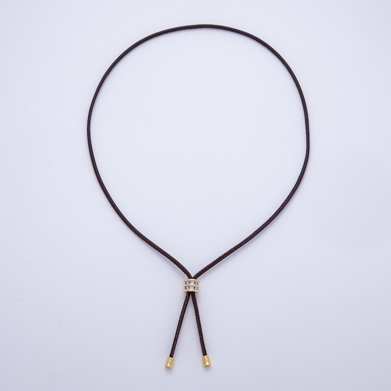 May include: A dark brown cord necklace with a gold-colored accent piece and gold-colored tips. The necklace is displayed against a white background. The cord forms a loop and then splits into two strands.