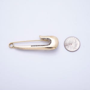 May include: A gold-colored safety pin with a row of small, dark stones along the curved end. The pin is next to a U.S. quarter for size comparison. The safety pin is a jewelry item.