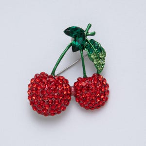 Puede incluir: Broche de cerezas con dos cerezas rojas con cristales, tallos y hojas verdes. El broche tiene un alfiler plateado. Las cerezas son de un rojo brillante, y las hojas son de varios tonos verdes.