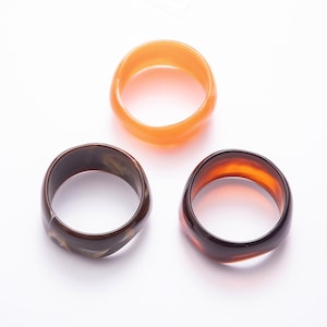 May include: Three circular rings made of smooth, polished resin. One ring is a translucent, solid orange color. The second ring features a dark brown and cream marbled pattern. The third ring is a deep, translucent reddish-brown color.
