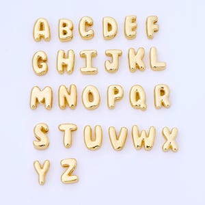 Puede incluir: Un conjunto de abalorios de letras del alfabeto doradas, cada uno con un diseño redondeado en forma de burbuja. Se muestran las letras de la A a la Z, perfectas para joyería personalizada o proyectos de manualidades. Las letras están dispuestas en filas.