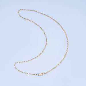 May include: A delicate gold-coloured necklace with a flat, elongated link chain design. The necklace is displayed against a light blue background, showcasing its subtle shine and intricate pattern. The clasp is visible at the bottom.