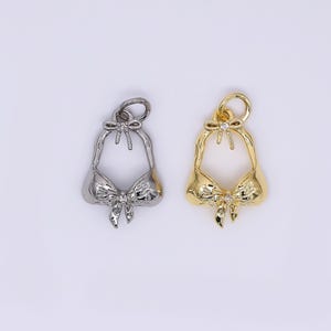 May include: Two metal bikini-shaped charms. One is silver-colored, the other is gold-colored. Both charms have a bow detail at the center and a small loop at the top for hanging. The charms are on a white background.