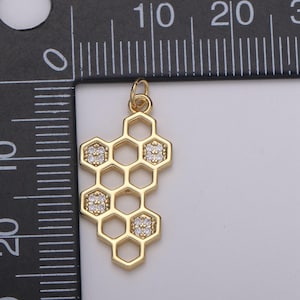 May include: A gold-colored pendant in the shape of a honeycomb. The pendant features a series of hexagonal cells, with four cells containing small, sparkling, clear gemstones. The pendant is designed for jewelry making.
