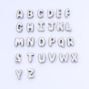 May include: A collection of silver-colored alphabet letter charms, each with a rounded, bubble-like design. The letters A through Z are displayed, perfect for jewelry making or crafting projects. The letters are arranged in neat rows against a white background.