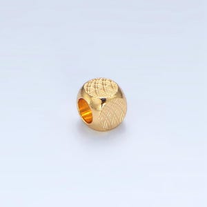May include: A gold-colored bead with a geometric pattern. The bead has a cylindrical shape with a hole running through the center. The surface is textured with a crosshatch design, adding visual interest to the jewelry component.
