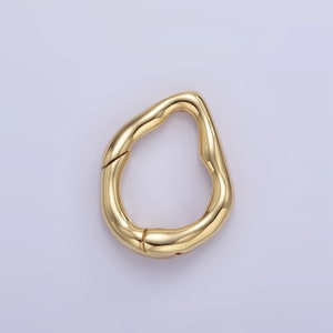 Puede incluir: Un cierre dorado de forma irregular. El cierre tiene una superficie lisa y reflectante y una abertura con bisagras. El diseño es orgánico y moderno, adecuado para joyas o accesorios. El cierre se muestra sobre un fondo morado claro.