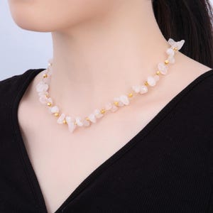 May include: A delicate necklace featuring small, irregular rose quartz stones interspersed with gold-colored beads. The necklace is strung on a thin, gold-colored wire and rests on a black top. The rose quartz has a soft pink hue.