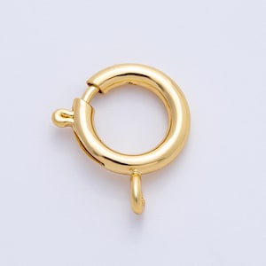 Puede incluir: Un cierre de anillo de resorte de color dorado, un componente de joyería, se muestra sobre un fondo blanco. El cierre es circular con una pequeña palanca y un lazo para sujetar a las piezas de joyería. El metal tiene una superficie brillante y reflectante.