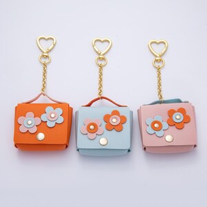 May include: Three small, square leather keychains in orange, light blue, and pink. Each keychain features a flower design and a gold-colored chain and heart-shaped clasp. The keychains have a small handle and a snap closure.