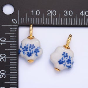 Op de afbeelding: Twee witte keramische bloemenbedels met blauwe bloemenmotieven en gouden accenten. De bedels zijn ongeveer 2,5 cm in diameter.