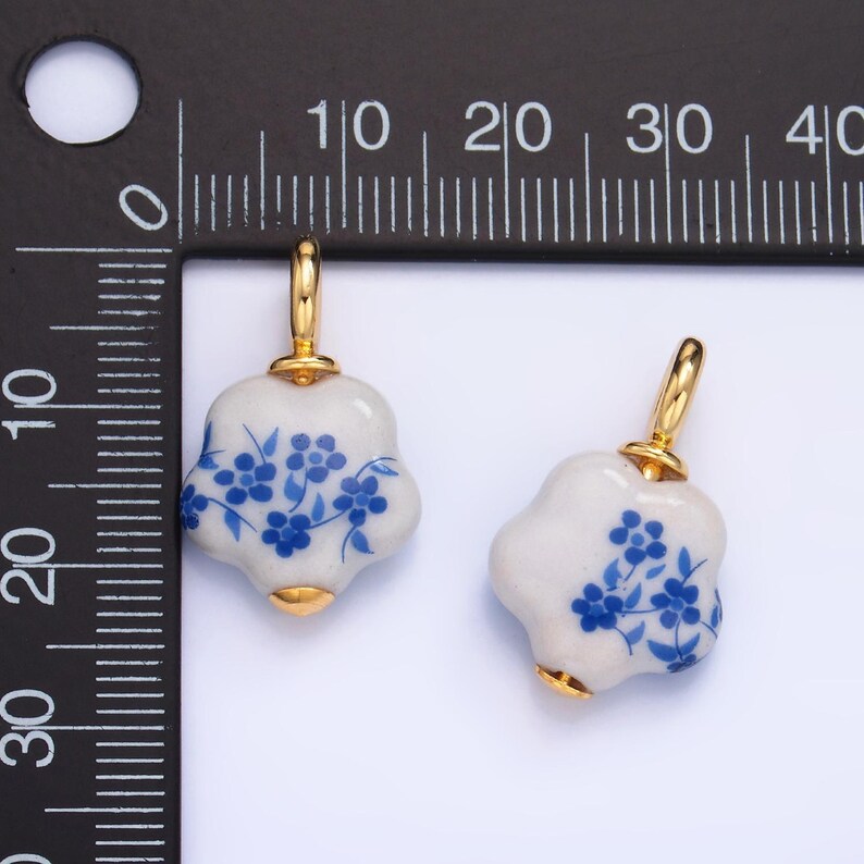 Op de afbeelding: Twee witte keramische bloemenbedels met blauwe bloemenmotieven en gouden accenten. De bedels zijn ongeveer 2,5 cm in diameter.