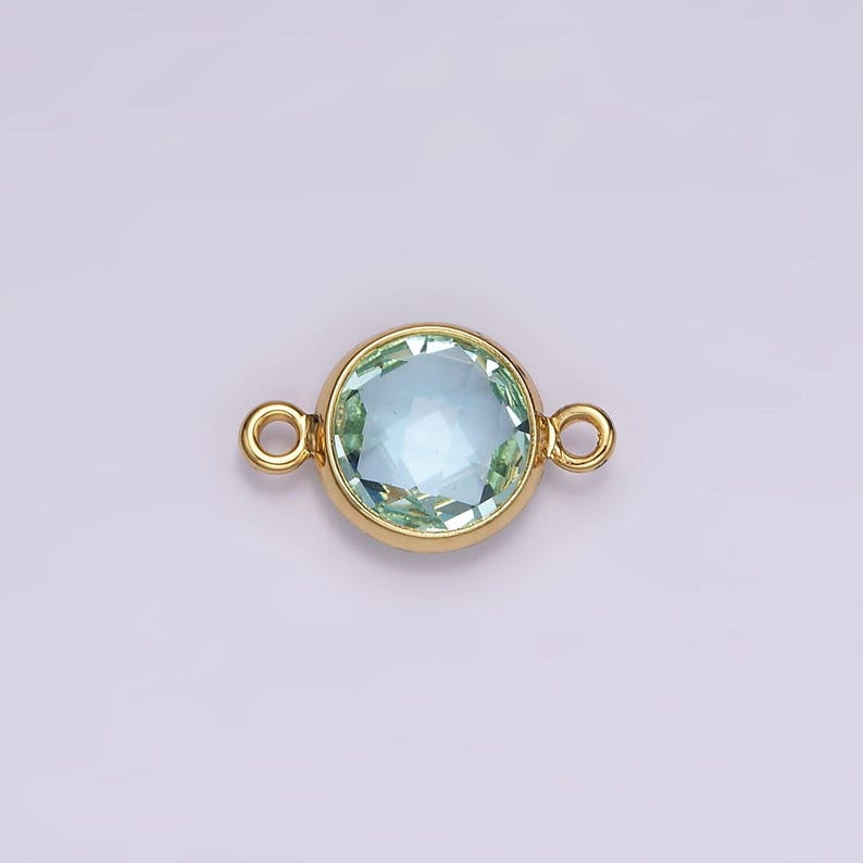 Puede incluir: Conector dorado con una piedra redonda de color azul claro. El conector tiene dos bucles a cada lado.