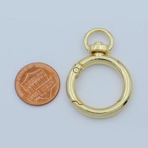 Puede incluir: Un cierre redondo de color dorado con un mecanismo de apertura accionado por resorte. El cierre tiene un peque&ntilde;o anillo en la parte superior y se muestra junto a una moneda de un centavo estadounidense para comparar el tama&ntilde;o. El cierre probablemente se utiliza para sujetar objetos.