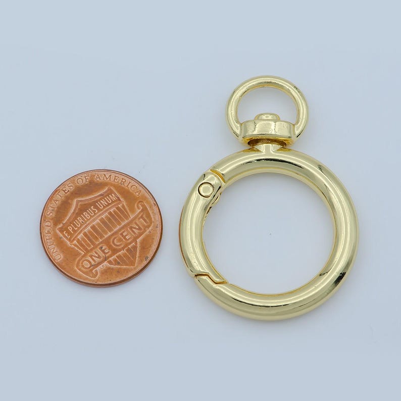 Puede incluir: Un cierre redondo de color dorado con un mecanismo de apertura accionado por resorte. El cierre tiene un peque&ntilde;o anillo en la parte superior y se muestra junto a una moneda de un centavo estadounidense para comparar el tama&ntilde;o. El cierre probablemente se utiliza para sujetar objetos.