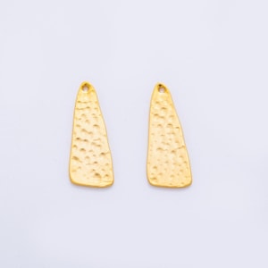 May include: Two gold-colored, hammered-texture metal pendants. Each pendant is a long, irregular triangle shape with a small hole at the top for hanging. The pendants are displayed against a white background.