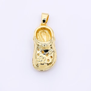 May include: A gold-colored pendant in the shape of a shoe, featuring a flower design on the front. The pendant has a loop at the top for a chain. The shoe has several holes and a textured interior.