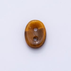 May include: An oval-shaped button with two holes. The button is a mix of brown and gold, with a polished, glossy finish. The button's design resembles tiger's eye stone, with natural patterns and color variations.