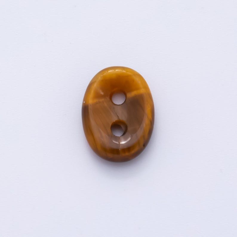 May include: An oval-shaped button with two holes. The button is a mix of brown and gold, with a polished, glossy finish. The button's design resembles tiger's eye stone, with natural patterns and color variations.