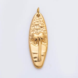 May include: Gold-colored pendant in the shape of a surfboard, featuring a palm tree and a beach scene. The pendant has a textured surface and a small loop at the top for a chain. The design is detailed and the overall aesthetic is tropical.