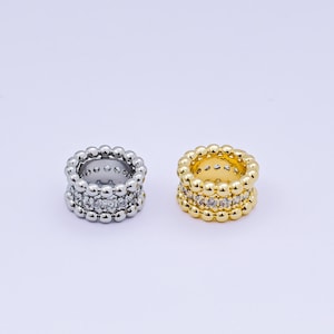 May include: Two rings, one silver and one gold, are displayed. The silver ring features a beaded design on the top and bottom, with a row of clear stones in the middle. The gold ring has a similar design, with clear stones in the center.