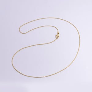 May include: A delicate gold-coloured necklace with a small clasp. The chain is thin and appears to be made of small, square links. The necklace is displayed on a white background.