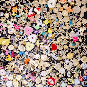 May include: A collection of gold-toned charms and pendants with various designs, including butterflies, hearts, and coins. Many charms are adorned with colorful enamel and gemstones. The assortment is arranged on a dark surface, creating a visually rich display.