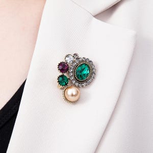 May include: A decorative brooch featuring a large central oval emerald green crystal surrounded by smaller clear crystals, a deep purple round crystal, a smaller emerald green round crystal, and a single smooth cream-colored faux pearl bead.