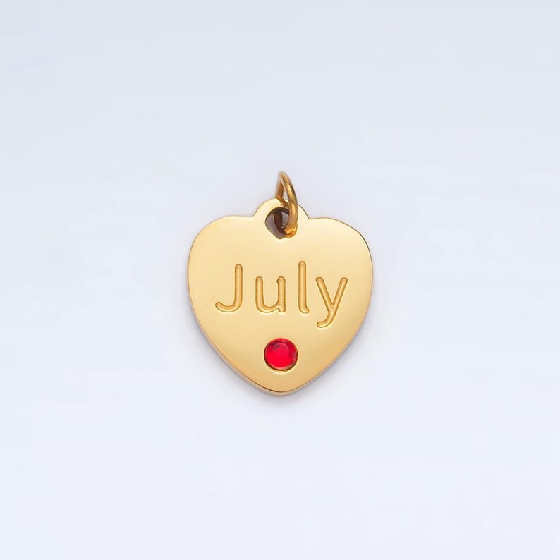 May include: Gold heart-shaped charm with the word "July" engraved on it. A small red gemstone is set in the center of the charm.
