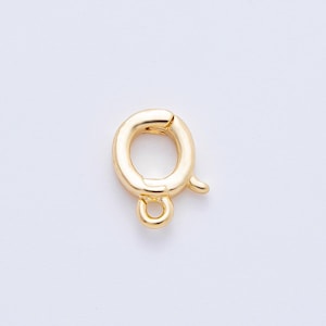 May include: A gold-colored spring clasp, a jewelry component, is shown against a white background. The clasp is oval-shaped with a small loop at the bottom and a curved lever on the side for opening and closing.