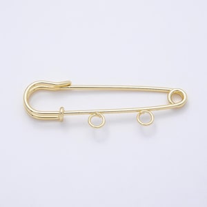 May include: A gold-toned metal safety pin featuring a smooth, polished finish. The pin includes a standard clasp mechanism on one end and two small, circular jump rings attached to the bottom bar, designed for hanging charms or decorative elements.