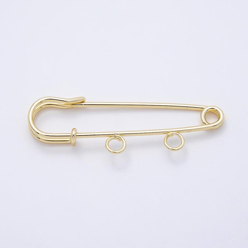 May include: A gold-toned metal safety pin featuring a smooth, polished finish. The pin includes a standard clasp mechanism on one end and two small, circular jump rings attached to the bottom bar, designed for hanging charms or decorative elements.