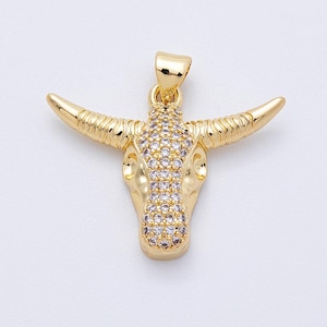 May include: A gold-colored pendant in the shape of a bull's head. The bull's head features a detailed design with horns and a face adorned with small, clear gemstones. The pendant has a loop at the top for a chain.