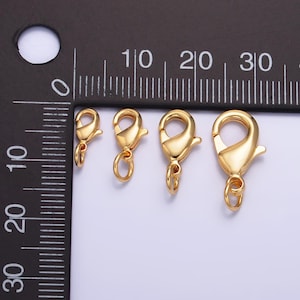 May include: Three gold-toned lobster clasps of different sizes. The clasps are shown against a black ruler with white markings.
