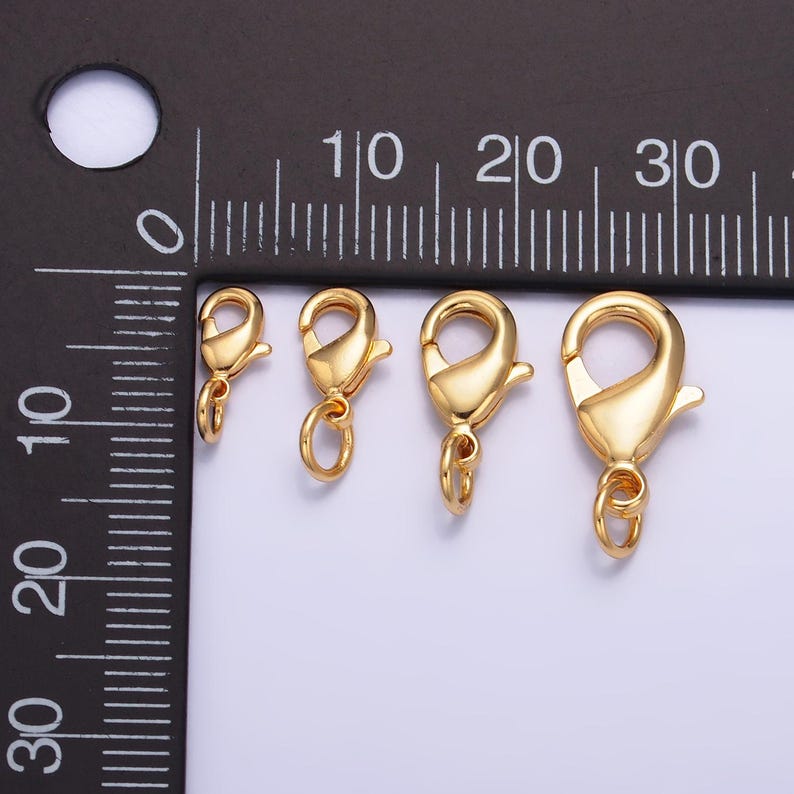 May include: Three gold-toned lobster clasps of different sizes. The clasps are shown against a black ruler with white markings.