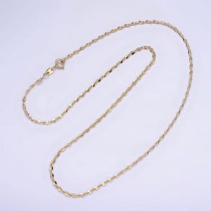 May include: A delicate gold-colored necklace with a repeating, flattened oval link design. The necklace is displayed on a white background, showcasing its intricate pattern and the warm tone of the metal. The clasp is visible at the top.