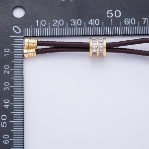 May include: A close-up of a brown leather cord necklace with gold-colored accents. The necklace features a gold-colored cylindrical bead with three rows of square, clear stones. The necklace is measured against a black ruler.