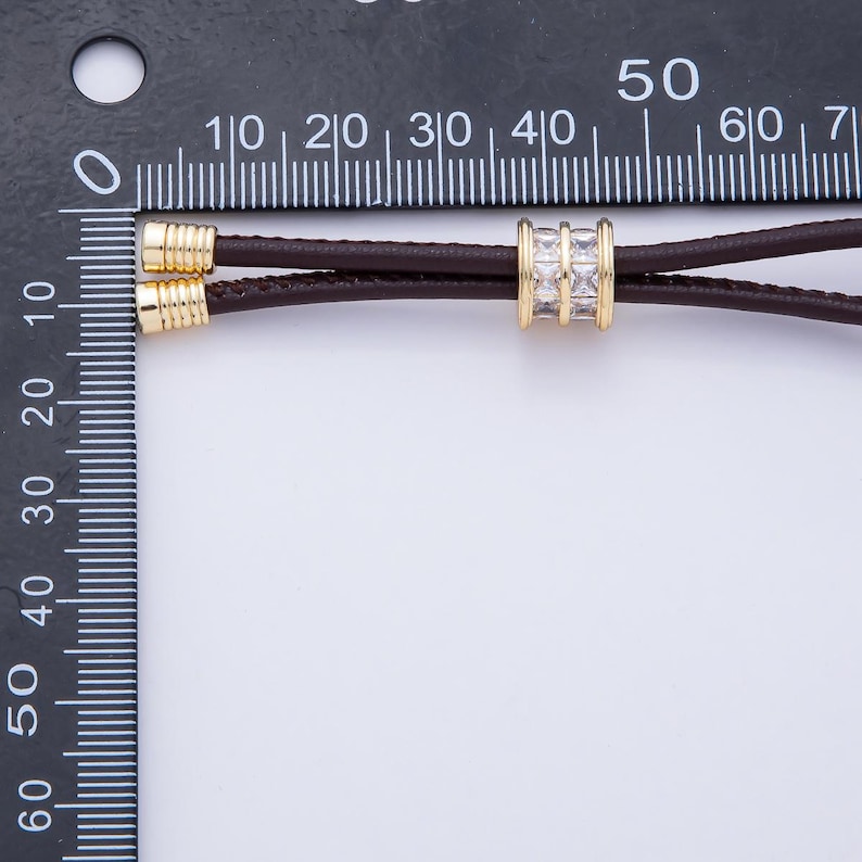 May include: A close-up of a brown leather cord necklace with gold-colored accents. The necklace features a gold-colored cylindrical bead with three rows of square, clear stones. The necklace is measured against a black ruler.