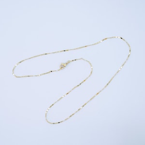 May include: A delicate gold-colored necklace with a small clasp. The chain is made of small, rectangular links that catch the light. The necklace is displayed on a white background, highlighting its simple elegance. A piece of jewelry.