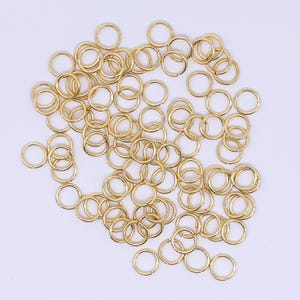 May include: A collection of small, gold-coloured jump rings scattered on a white surface. The rings are circular and appear to be made of metal. They are likely used for jewellery making or other craft projects.