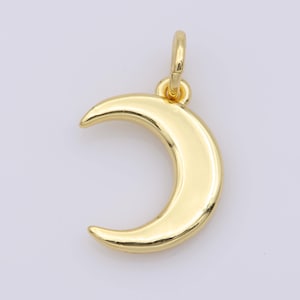 Puede incluir: Un colgante de luna creciente de color dorado, con un lazo para sujetarlo a una cadena. El colgante tiene una superficie lisa y pulida y una forma curva. La joya es probablemente de metal.