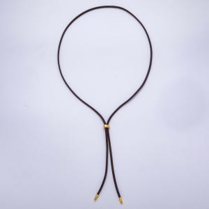 K&ouml;nnte beinhalten: Eine dunkelbraune Halskette mit einem goldfarbenen Metallakzent. Die Halskette hat ein kreisf&ouml;rmiges Design mit zwei herabh&auml;ngenden Kordeln, die in goldfarbenen Spitzen enden. Der goldfarbene Akzent ist ein kleines, rundes Band.