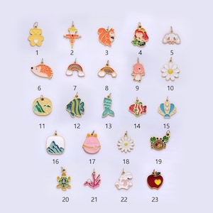 Puede incluir: Veintitrés abalorios de metal dorado con diseños de esmalte de colores. Los abalorios presentan varios animales, objetos y símbolos, incluyendo un oso, un espantapájaros, una ardilla, una sirena, un pez, una montaña, un arcoíris, una margarita, una tortuga y una manzana.