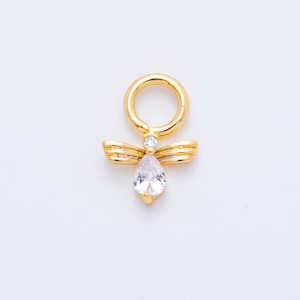 Puede incluir: Un dije dorado con un pequeño anillo en la parte superior. El dije presenta un par de alas estilizadas y una piedra preciosa clara en forma de pera. Una piedra preciosa redonda y clara más pequeña está engastada encima de la forma de pera.