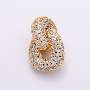 May include: A gold-toned pendant featuring two interlocking oval links. The surface of both links is densely encrusted with numerous small, clear, round-cut cubic zirconia stones that create a highly reflective and sparkling appearance.