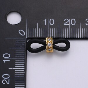 Puede incluir: Un amuleto de oro infinito con diamantes de imitaci&oacute;n transparentes engastados en una banda de goma negra. El amuleto tiene aproximadamente 1 cm de di&aacute;metro.