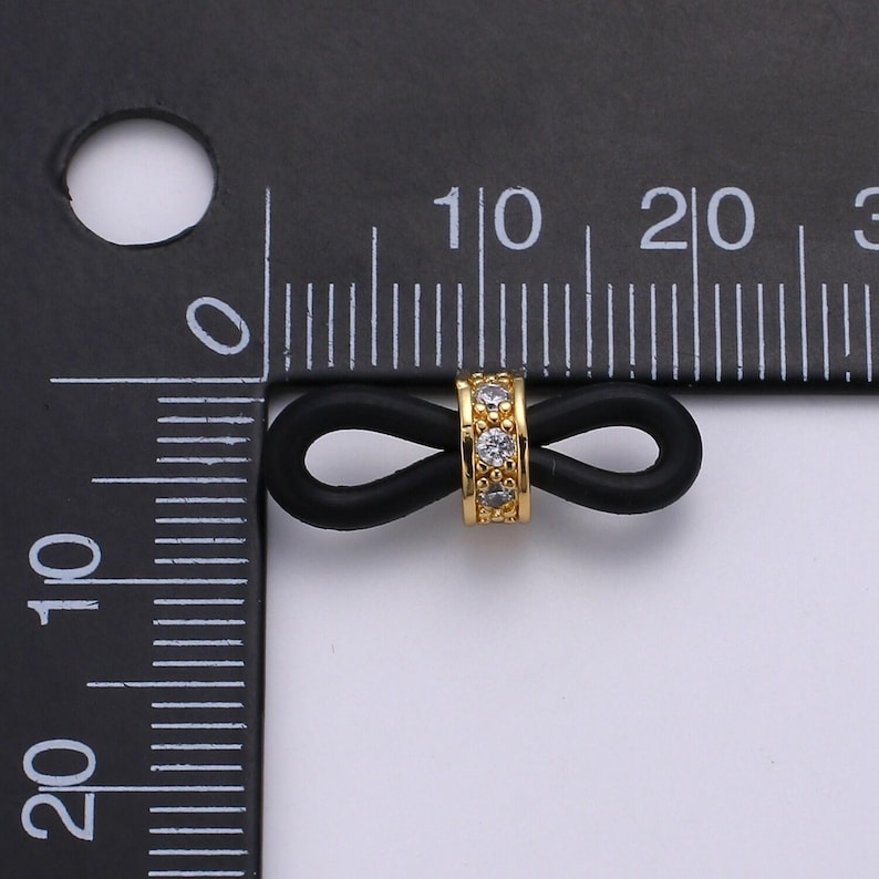 Puede incluir: Un amuleto de oro infinito con diamantes de imitaci&oacute;n transparentes engastados en una banda de goma negra. El amuleto tiene aproximadamente 1 cm de di&aacute;metro.