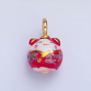 May include: A red and white ceramic charm in the shape of a smiling cat wearing a red and gold patterned kimono. The charm has a gold loop for hanging.